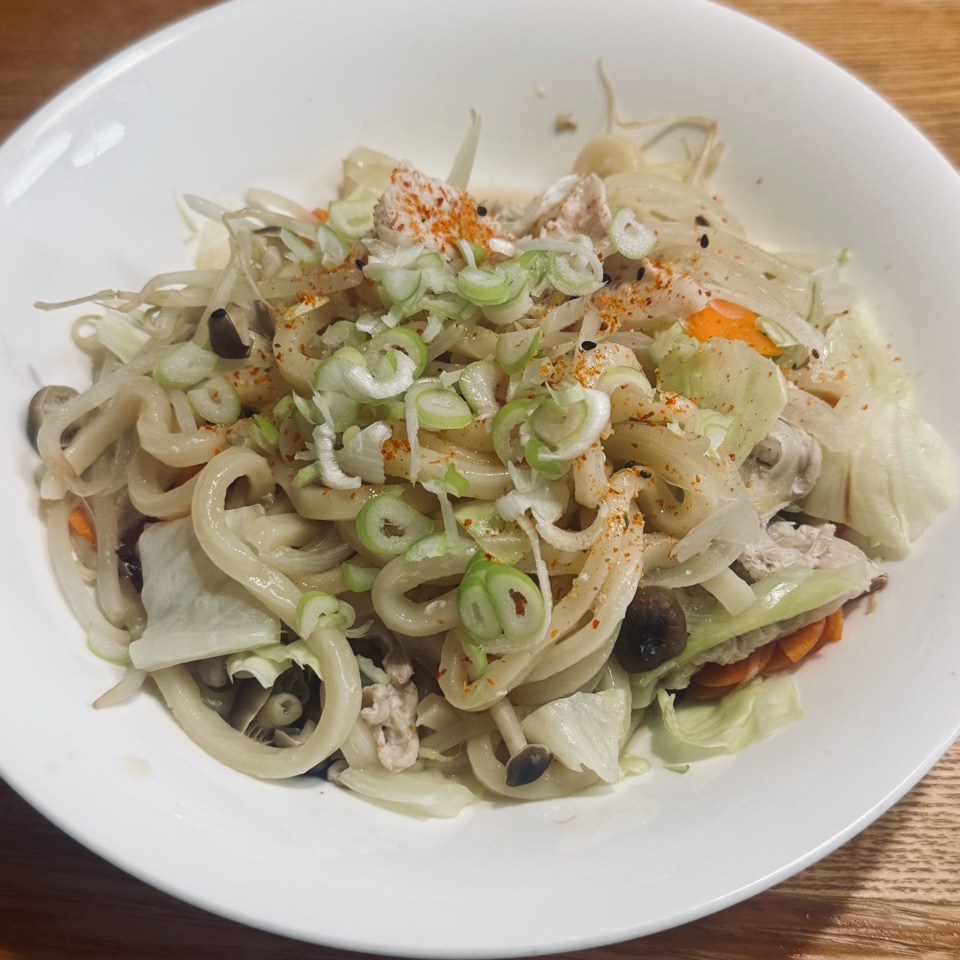 白い皿に盛ったレンチン蒸しうどん。うどん、キャベツ、にんじん、しめじ、豚肉に小口ねぎと七味をかけたもの